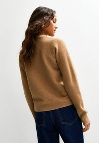 Woman with long wavy hair wears a camel-colored sweater and dark blue jeans, facing sideways against a plain light background.