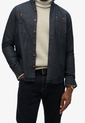 Mand iført mørk denim skjorte med knapper over cremefarvet strikket rullekravetrøje, mørke bukser, sort bælte og guldfarvet armbåndsur med læderrem.