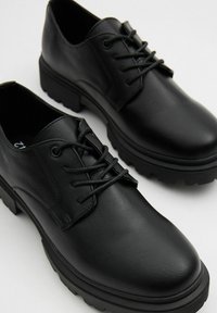 Zapatos de cuero negro con un acabado suave, punta redonda y una suela de goma gruesa y texturizada. Presentan cordones y acentos de diseño minimalista.