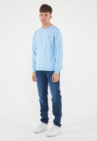 Maglione azzurro chiaro a maniche lunghe con colletto rotondo e polsini a coste, abbinato a jeans blu scuri e sneakers bianche. Logo semplice sul petto.