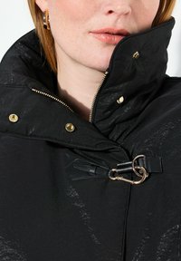 Nahaufnahme einer Person, die eine schwarze Jacke mit goldenem Reißverschluss, Druckknöpfen und Verschluss trägt. Man sieht die Lippen und einen Teil des Gesichts sowie des Ohrs mit Ohrhängern.
