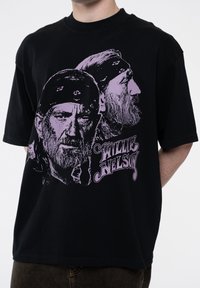 Black cotton T-shirt featuring purple graphic illustrations of two side profiles of a man with bandanas, with "WILLIE NELSON" text below.