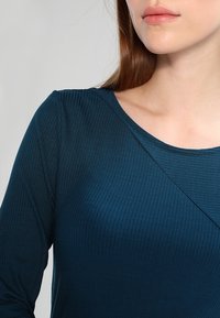 Top a maniche lunghe ribaltato in teal con scollatura rotonda e dettaglio della cucitura diagonale sul petto. Texture morbida e silhouette aderente.