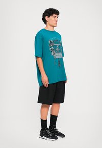 Fiatal férfi oldalról állva, türkiz színű, oversized madárkalitka mintás pólót visel, fekete rövidnadrággal, fekete zoknival és fekete sportcipővel.