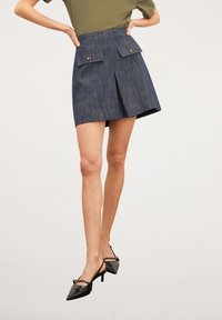 Jupe en denim bleu foncé avec pli à l'avant, deux poches à rabat boutonnées et taille cintrée. Associée à des escarpins à bride arrière noirs à bout pointu.