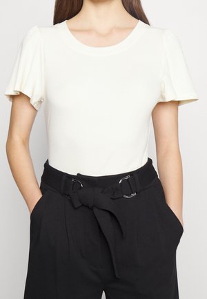Kobieta ubrana w białą bluzkę z krótkim rękawem z bufkami oraz czarne spodnie z wysokim stanem, przewiązane materiałowym paskiem.