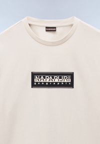 Korte mouwen crèmekleurig katoen t-shirt met een zwart rechthoekig logo gecentreerd op de borst met de tekst "NAPAPIJRI geographic" in het wit.