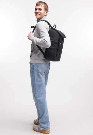 Sac à dos noir en tissu texturé avec une fermeture enroulable, des bretelles rembourrées et une poche latérale visible. Modèle portant des vêtements décontractés.