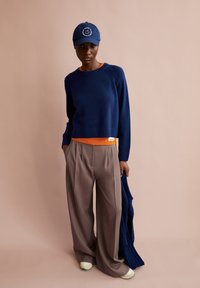 Navy sweater layered over orange top, wide beige trousers, and white sneakers. Blue baseball cap with a logo, textured fabric, relaxed fit.