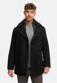 Schwarzer doppelt geknöpfter Mantel mit breiten Revers, Knopfschluss und seitlichen Taschen. Getragen über einem hellgrauen gerippten Pullover.