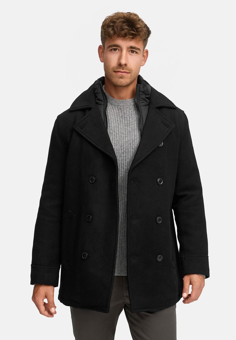 Schwarzer doppelt geknöpfter Mantel mit breiten Revers, Knopfschluss und seitlichen Taschen. Getragen über einem hellgrauen gerippten Pullover.