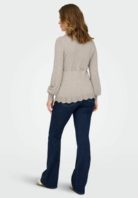 Svetlo sivi pleteni pulover z zvonastimi rokavi, valovitim robom in tremi gumbi na hrbtu, v kombinaciji s temno modrimi razširjenimi kavbojkami.