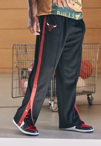 Crne sportske hlače s crvenim bočnim prugama, s vezom loga bika. Uparene s crnim i crvenim tenisicama koje imaju slip-on dizajn.