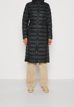 Persoon draagt een lange zwarte gewatteerde pufferjas met capuchon, beige broek met wijde pijpen en beige instap schoenen, staand op een witte achtergrond.