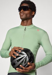 Man in light green zip-up jersey holding a black bike helmet, wearing large dark sunglasses, against a plain light background.