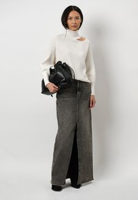 Crème, geribbelde trui met een hoge hals en een zijuitsparing. Lange, grijze denimschoot met een voorkniplit. Model houdt een zwarte, geplooide handtas vast.