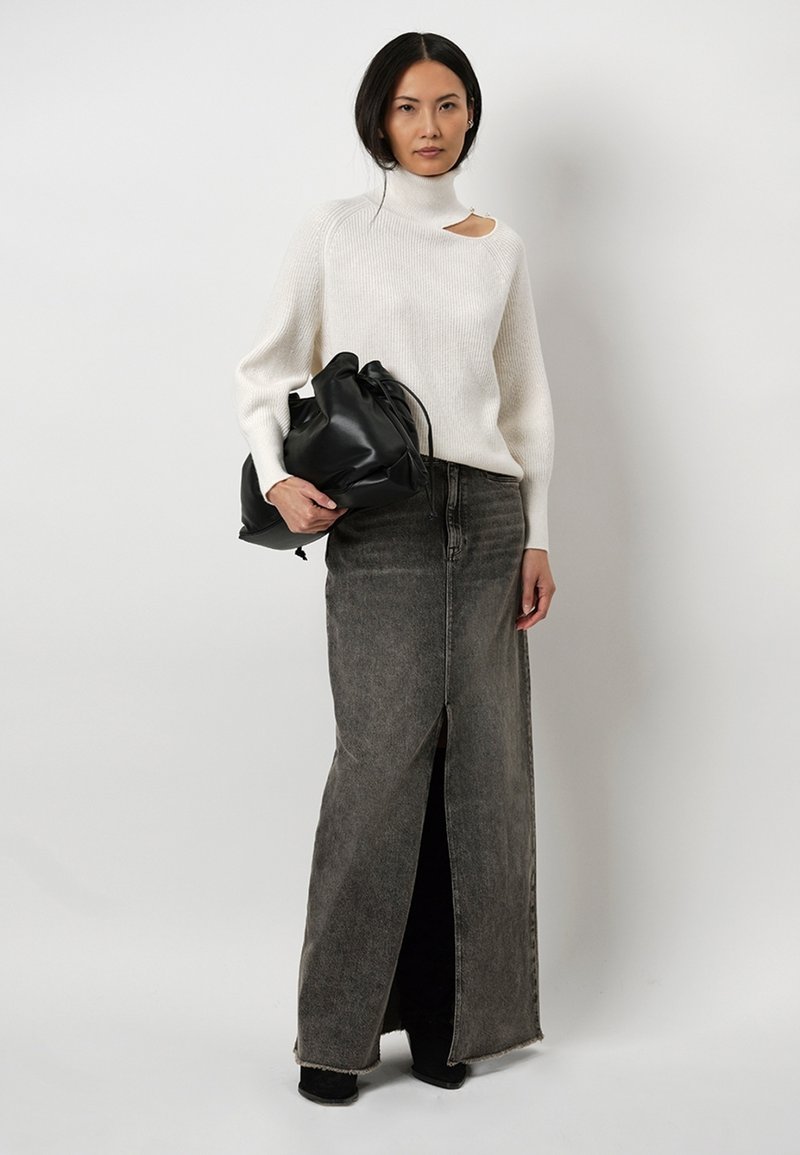Crème, geribbelde trui met een hoge hals en een zijuitsparing. Lange, grijze denimschoot met een voorkniplit. Model houdt een zwarte, geplooide handtas vast.