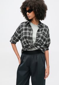 Camisa de flanela xadrez preta e cinzenta amarrada na cintura sobre uma t-shirt cinzenta clara, combinada com calças escuras de cintura alta. Acessórios incluem óculos de sol redondos pretos.