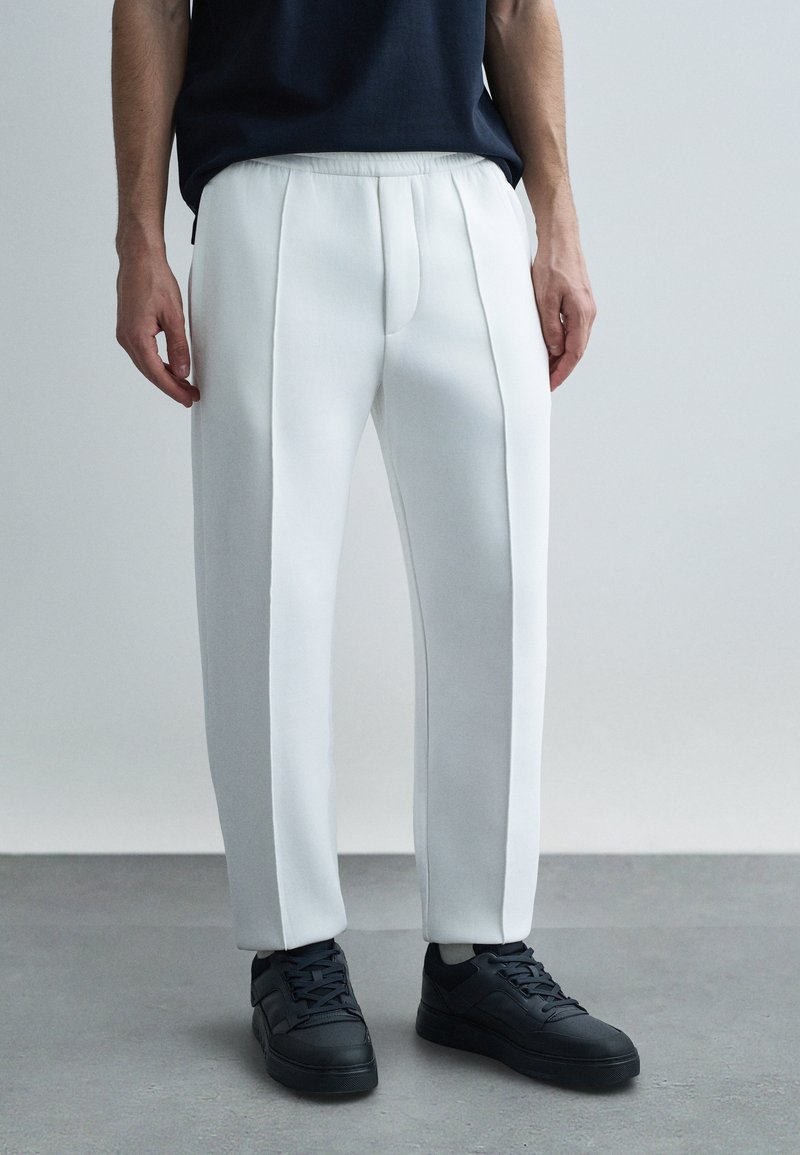 Biele nohavice na mieru so voľným strihom, bočnými záhybmi a elastickým pásom. Kombinované s čiernymi teniskami pre kontrast.