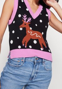 Gilet sans manches en tricot noir avec bordure rose, pois blancs et un renne marron aux bois roses, porté avec un jean bleu.