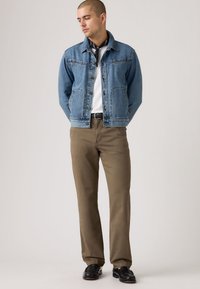 Τζιν μπουφάν σε ανοιχτό μπλε χρώμα, με κουμπιά και τσέπες στο στήθος, συνδυασμένο με ανοιχτό καφέ παντελόνι και μαύρα παπούτσια, φορεμένο πάνω από λευκό T-shirt.