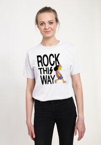 Žena v bílém tričku s nápisem "ROCK THIS WAY" a kreslenou postavičkou s sluchátky, která kráčí, v kombinaci s černými kalhotami.