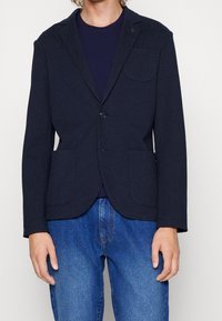 Marinblå bomullskavaj med spetsig krage, två framfickor, enkel knappstängning och en bröstficka, bärs över en marinblå skjorta.