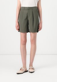 Shorts taillés en khaki à taille haute, avec des plis à l'avant et une coupe droite. Associés à des ballerines beiges ornées d'une sangle et d'une boucle.