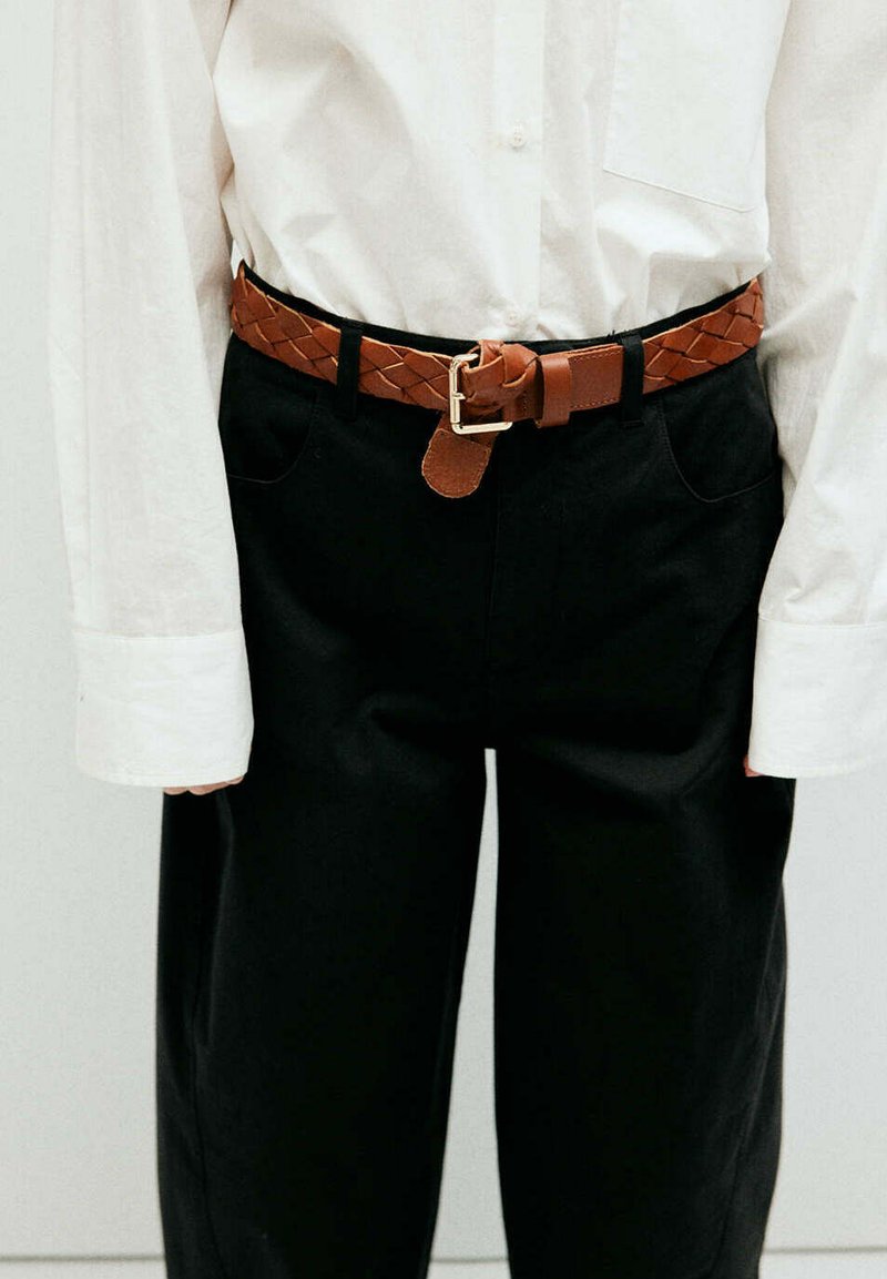 Personne portant une chemise blanche à manches longues rentrée dans un pantalon noir avec une ceinture en cuir tressé marron attachée à la taille.