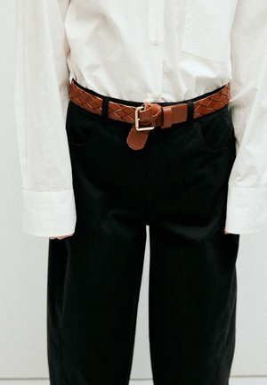 Persona que lleva una camisa blanca de manga larga metida en pantalones negros con un cinturón de cuero trenzado marrón abrochado en la cintura.
