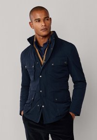 Mand iført en marineblå quiltet jakke med fire lommer foran, lagdelt over en tan sweater og en mørk skjorte, stående med hænderne i lommerne.