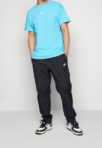 Persona vestida con una camiseta ligera azul de Nike, pantalones cargo negros de Nike y zapatillas negras y blancas de Nike, de pie con las manos en los bolsillos.
