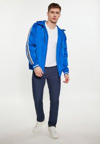 Giacca leggera blu con strisce bianche e grigie sulle maniche, abbinata a jeans scuri e sneakers bianche. Tessuto liscio, vestibilità casual.
