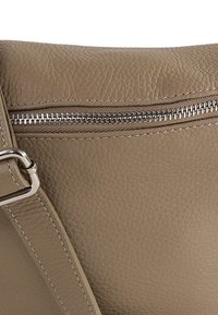 Beige leren crossbodytas met een gestructureerd oppervlak, voorzien van een zilveren ritssluiting en banddetail. De stiksels hebben dezelfde kleur.