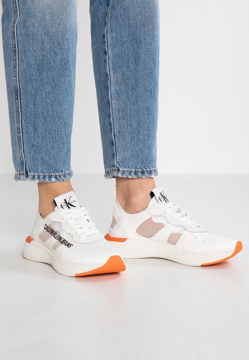 White Calvin Klein mesh sneakers with orange soles worn with blue jeans, shown from side and front angles on a smooth floor.
