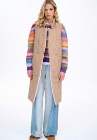 Beżowa, pikowana kamizelka bez rękawów z wysokim kołnierzem, z dwoma guzikami. Nosi się ją na kolorowy sweter w paski oraz dżinsowe spodnie w stylu wide-leg.