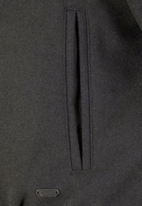 Dark gray wool coat featuring a vertical pocket and a metal designer label. The fabric has a soft texture and a clean finish.