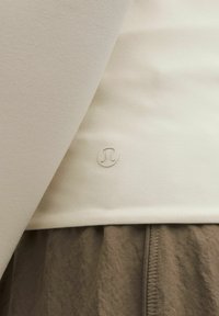 Zatvoren kadar bijele odjeće s malim Lululemon logotipom blizu rubova, u kombinaciji s smešnim teksturiranim hlačama.