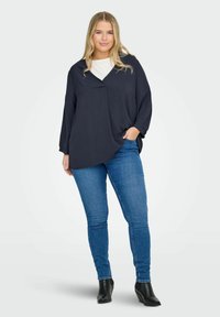 Tmavě navy volná blůza s výstřihem do V a pokleslými rameny, v kombinaci s modrými skinny džíny a černými kotníkovými botami. Hladká textura látky.