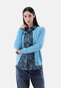 Hellblaues Strickcardigan mit rundem Ausschnitt, Knopfschluss und gerippten Bündchen über einer dunkel gemusterten Bluse mit Herzen. Trägt dazu eine Jeans.