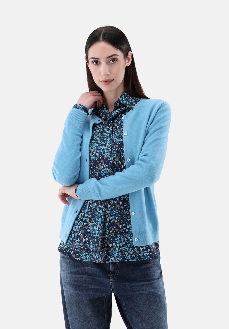 Hellblaues Strickcardigan mit rundem Ausschnitt, Knopfschluss und gerippten Bündchen über einer dunkel gemusterten Bluse mit Herzen. Trägt dazu eine Jeans.