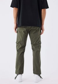 Persoon die olijfgroene cargobroek, zwart T-shirt en witte sneakers draagt, met de rug naar de kijker toe staand tegen een effen achtergrond.