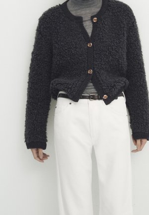 Person wearing black textured buttoned cardigan over gray top paired with white high-waisted pants and black belt against white background.