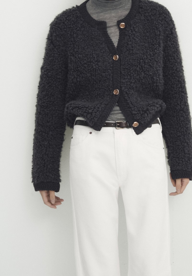 Personne portant un cardigan noir texturé à boutons par-dessus un haut gris, associé à un pantalon blanc taille haute et une ceinture noire, sur fond blanc.