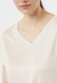 Maglietta a V-collo color crema realizzata in un tessuto morbido e leggero, con maniche corte e una leggera lucentezza, caratterizzata da un design semplice e pulito.