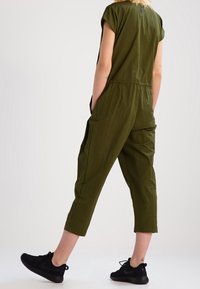 Jumpsuit verde oliva hecho de una tela ligera, con un corte holgado, mangas cortas, tobillos ajustados y bolsillos laterales. Combinado con zapatillas negras.