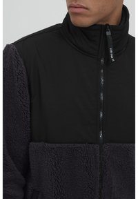 Svart jakke med glidelås, som har et mykt, teksturert mørkegrått fleece-materiale på kroppen og en glatt svart øvre del, med høy krage og merkevaredetalj.