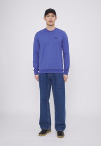 Felpa blu a maniche lunghe con collo rotondo e logo, abbinata a jeans blu a gamba larga e scarpe nere con suola color cammello.