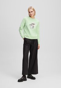 Felpa verde chiaro con stampa grafica di un gatto e di un personaggio, abbinata a pantaloni larghi neri e scarpe nere. Tessuto morbido, vestibilità rilassata.