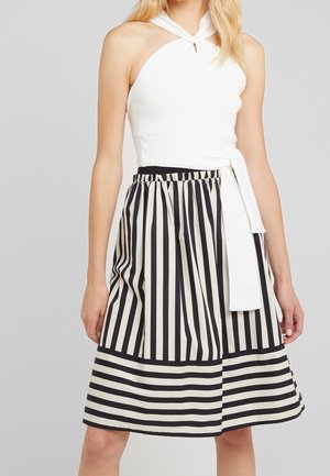 Mujer vistiendo una blusa blanca sin mangas con detalle de lazada en el cuello y una falda a la rodilla con rayas verticales y horizontales en blanco y negro.